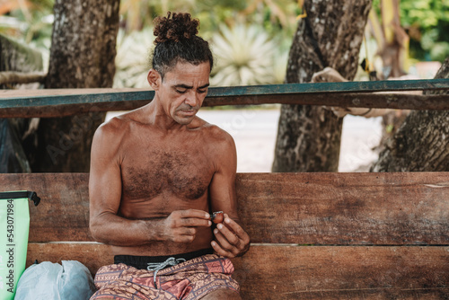 Imagen horizontal de un hombre adulto de cabello rizado sin camisa en la playa encendiendo su pipa para fumar hierba. 