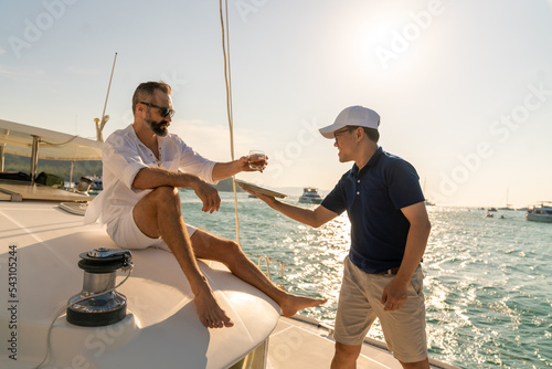 Asian man waiter serving alcohol drink to passenger tourist while catamaran boat yacht sailing in the ocean at summer sunset. Handsome man enjoy luxury outdoor lifestyle on holiday travel vacation.