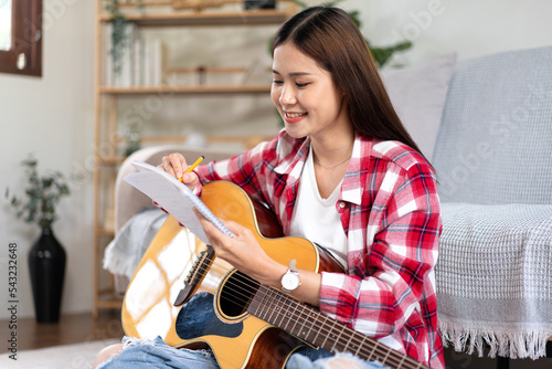 Young woman is compose song and writing lyrics on notebook while putting guitar on the legs and sitting