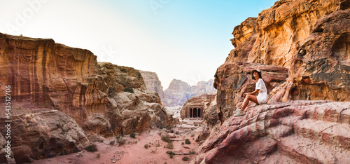 Caucasian young female tourist sit and smile while looking to beautiful Petra ancient city carvings. Famous visit Jordan attraction destination. Petra archeological site panorama