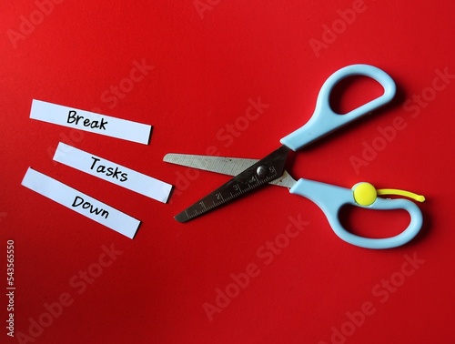 Scissors on red background, cut paper pieces with handwritten text - Break Tasks Down - means to break down tasks into smaller parts which easier to work on, focus on doing one work at a time