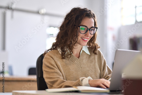 Young happy professional business woman worker employee sitting at desk working on laptop in corporate office. Smiling female student using computer technology learning online, doing web research.