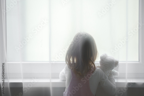 Little girl holding white teddy bear standing alone at window behind transparent day curtains and looking out from home. Rear view. Waiting concept. Close up.