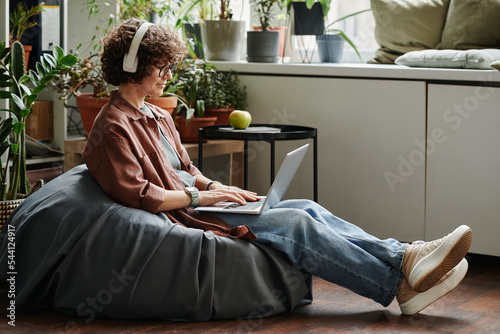 Young female employee in headphones and casualwear listening to music and networking while sitting in soft bag chair in modern office