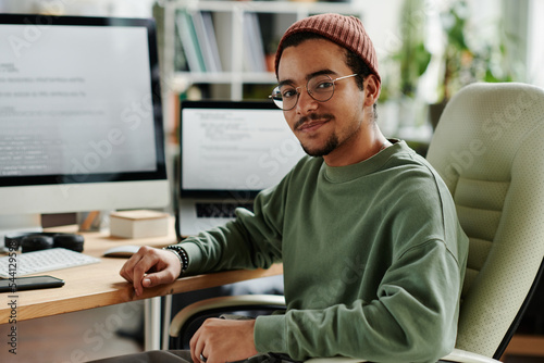 Young confident IT engineer or software developer in casualwear sitting in armchair by computer monitor and laptop and looking at camera