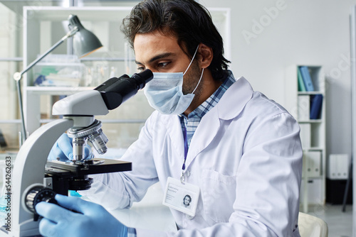 Young scientist in protective workwear looking in microscope during trial or creating effective vaccine against new dangerous virus