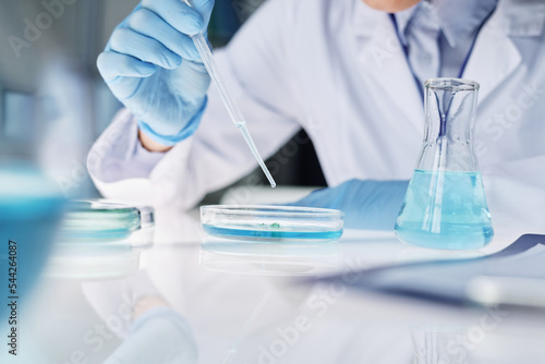 Close-up of clinician in gloves and labcoat dropping liquid substance in petri dish with bacteria or new virus during clinical experiment