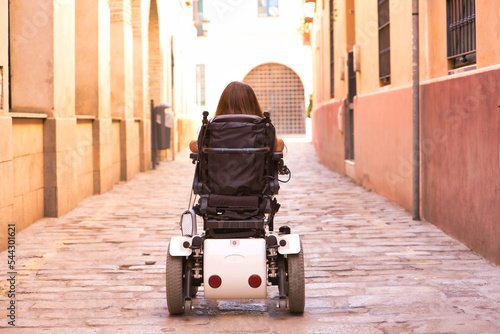 Disabled woman with reduced mobility and small stature in an electric wheelchair walking along a lonely city street, seen from the back. Concept handicap, disability, incapacity, special needs.