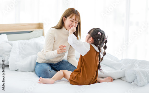 Closeup shot of Millennial Asian young female teenager mother nanny babysitter in casual sweater and jeans sitting eating snack feeding from little cute preschooler daughter girl on bed in bedroom