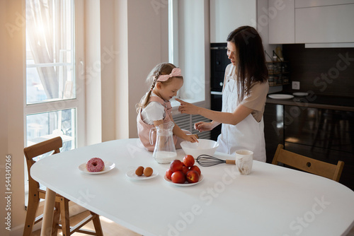 An image of a young woman and her little cute daughter are cooking in the kitchen. Have fun together, interfere with flour. Mom teaches child to cook