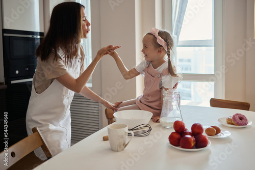 An image of a young woman and her little cute daughter are cooking in the kitchen. Have fun together, interfere with flour. Mom teaches child to cook