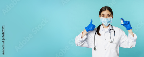 Portrait of smiling doctor, medical personel in face medical mask and rubber gloves, showing thumbs up and coronavirus, omicron vaccine, standing over blue background