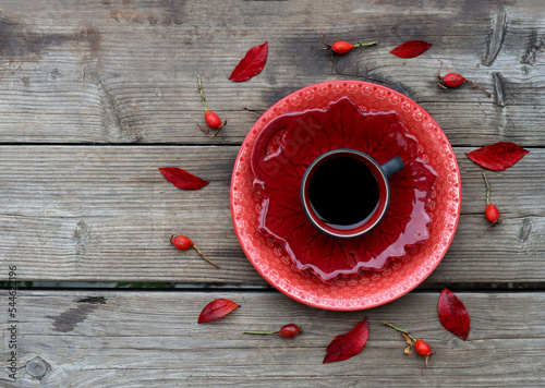 En kopp svart kaffe, koppen står på två röda tallrikar. Runt om finns röda löv och nypon. Bilden är tagen på trä. 