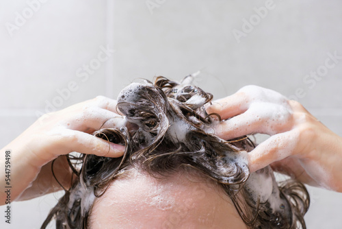 A girl washes her hair with shampoo on gray tiles background, front view.