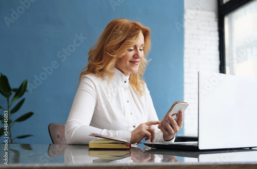 Beautiful business woman blonde in a blouse sits at a desk in front of an open laptop, holds a mobile phone in her hands and answers messages
