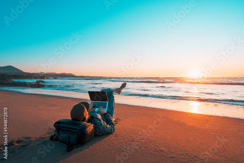 mature latin digital nomad laying on the sand of the beach with a laptop working at sunset over the ocean
