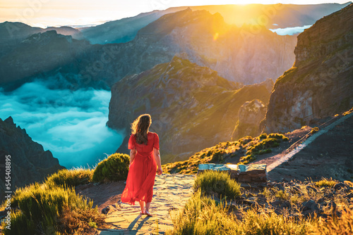 Beautiful woman in red dress walking barefoot on a very scenic hiking trail in the evening sun on Pico do Ariero. Verade do Pico Ruivo, Madeira Island, Portugal, Europe.