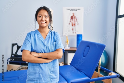 Young chinese woman wearing physiotherapist uniform standing with arms crossed gesture at rehab clinic