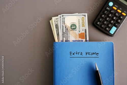 Cash dollar money, pen, calculator on a blue notebook with handwritten text on cover EXTRA INCOME on grey background, concept of make more money, additional income earn from part time side hustle job