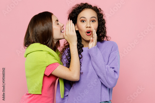 Young two friends shocked women wears green purple shirts together whispering gossip and tells secret behind her hand sharing news isolated on pastel plain light pink color background studio portrait.