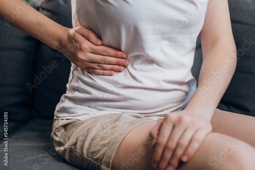 Close up of young female with fatty liver touches right side with hand, suffering from abdominal pain sit on grey couch. Pain in right side, appendix, gallstones and gynecological diseases concept