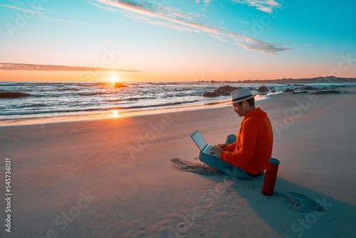 latino digital nomad sitting on the shore of the beach working at a beautiful sunset over the ocean.