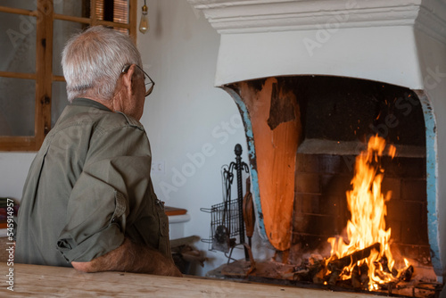 Hombre mirando el fuego en casa rústica vestido con ropa de camuflaje, cazador anciano de la tercera edad blanco y con canas un poco de espaldas