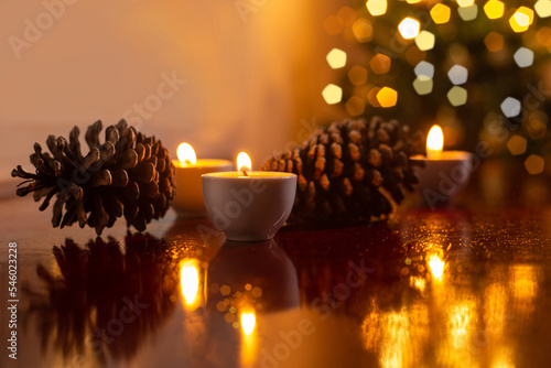 Decoração de Natal. Algumas velas acesas em uma superfície reflexiva, duas pinhas e um detalhe desfocado da árvore de Natal ao fundo.