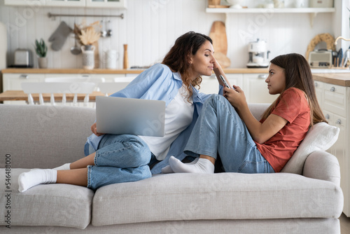 Curious unceremonious woman following teenage girl trying to read SMS message in child phone. Embarrassed schoolgirl sits on sofa in living room looks at mother who interferes in personal space