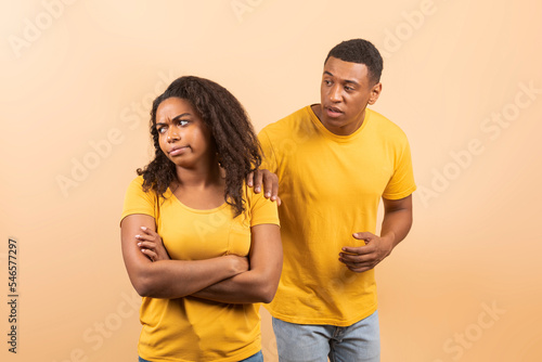 Black couple after quarrel, husband trying to talk and reconcile with offended wife, standing over peach background