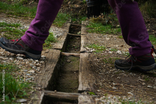 Foto della parte bassa di un escursionista che scavalca una canaletta di scolo in legno, lungo un sentiero in montagna