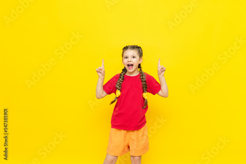 A little girl points her finger at an empty space for your advertisement on a yellow isolated background. A beautiful child is smiling in a red T-shirt with two pigtails. Copy space.
