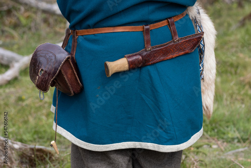 Back view of a viking man in blue wool tunic with sax knife in a leather sheath and a small leather bag on the belt.