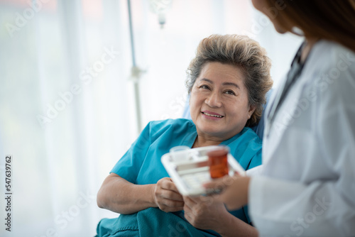 Smiling young doctor and nurse in lab coat with stethoscope giving care and medicinal syrup and treatment to senior woman resting on hospital bed while admitted for treatment against illness