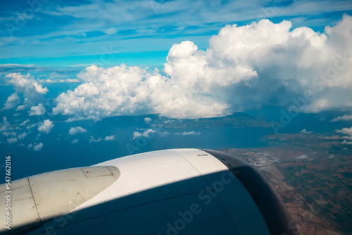 View from Boeing 737 MAX 8 on White Air Clouds, Clear Blue Sky And Surface Of Earth With Fields And Mountains. In frame White-Grey Engine Casing With Vanes.