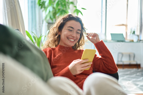 Happy relaxed young woman sitting on couch using cell phone, smiling lady laughing holding smartphone, looking at cellphone enjoying doing online ecommerce shopping in mobile apps or watching videos.