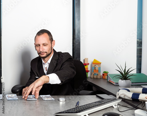Serious jewish man in formal wear sit at desk wearing kippah with surprised looking at dollar bills. Caucasian man is putting hundred dollar bills on the table as payment. Jew receiving salary money