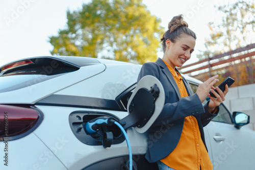 Young woman with smartphone waiting while her electric car charging in home charging station, sustainable and economic transportation concept.