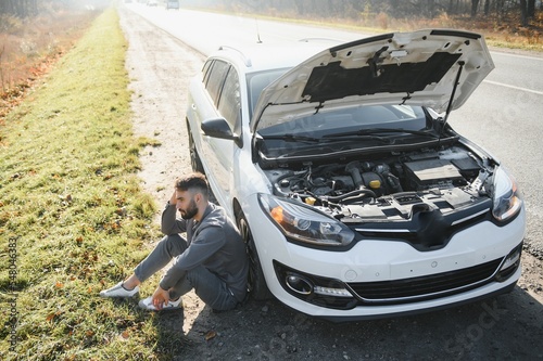 Picture of frustrated man sitting next to broken car with open hood