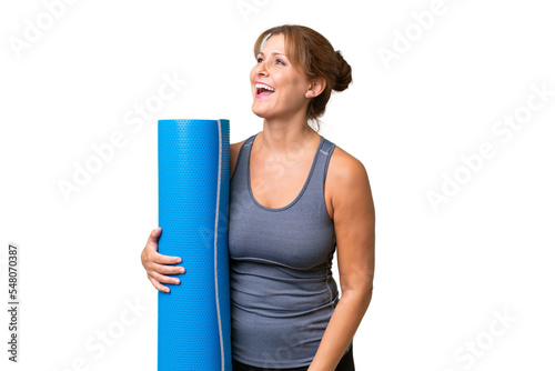 Middle-aged sport woman going to yoga classes while holding a mat over isolated background laughing in lateral position