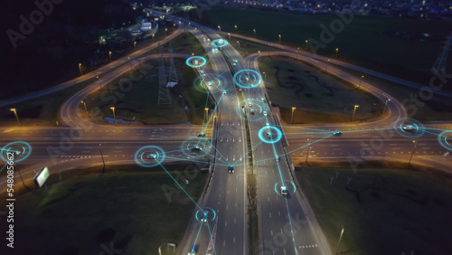 Self-driving autonomous cars move along a suburban traffic intersection in the evening. Neon HUD elements visualize the interaction of driverless cars connected to a common network