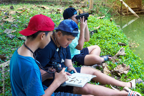 Asian boy and friends Invite each other to see birds in the community forest on holidays. And help each other to search for the types of birds found from the birds Guide book.