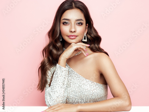 Beautiful woman in cocktail dress posing on pink background. Beauty model with long curly hair. Christmas or New Year festivities. Jewelry and manicure