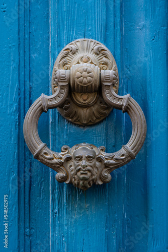 Closeup view of traditional vintage brass door knocker with face of man isolated on bright blue wooden door, Montpellier, France