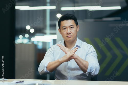 A serious young man Asian office worker sits at the desk in the office, crossed his arms in front of him, shows prostration, objection to the camera.