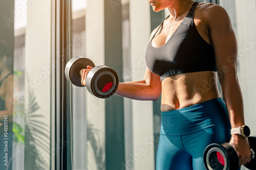 Asian woman standing lifting dumbbell exercise to increase arm muscles at gym. She wearing a black sports bra and blue pants.Fitness,Sport and healthy style concept.