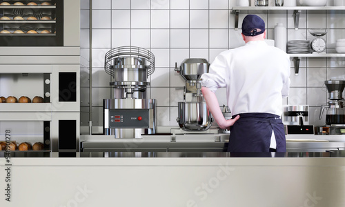 Male professional baker working in industrial bakery kitchen with kneading machines, equipments, bread dough in the manufacturing oven with empty stainless steel counter for product display