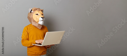 Portrait of busy man with lion mask with wearing casual style orange jumper posing isolated over gray background with copy space for advertisement, working on laptop.