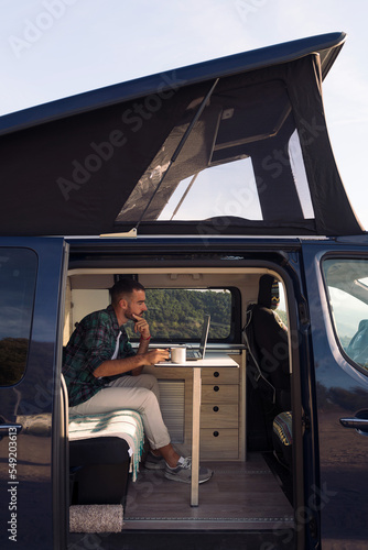 young man sitting in his camper van working on his laptop, concept of freedom and digital nomad lifestyle