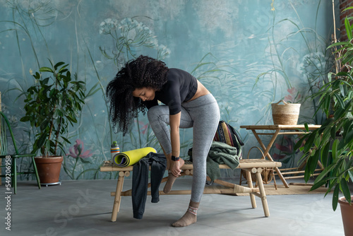 Curly African-American woman putting on clothes after fitness training over green domestic house plant loft blue home background. Lifestyle concept. Copy space, mock-up. Eco natural, body conscious
